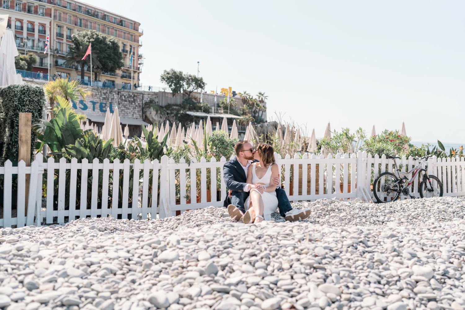 man and woman kiss sitting on rocky beach next to white picket fence