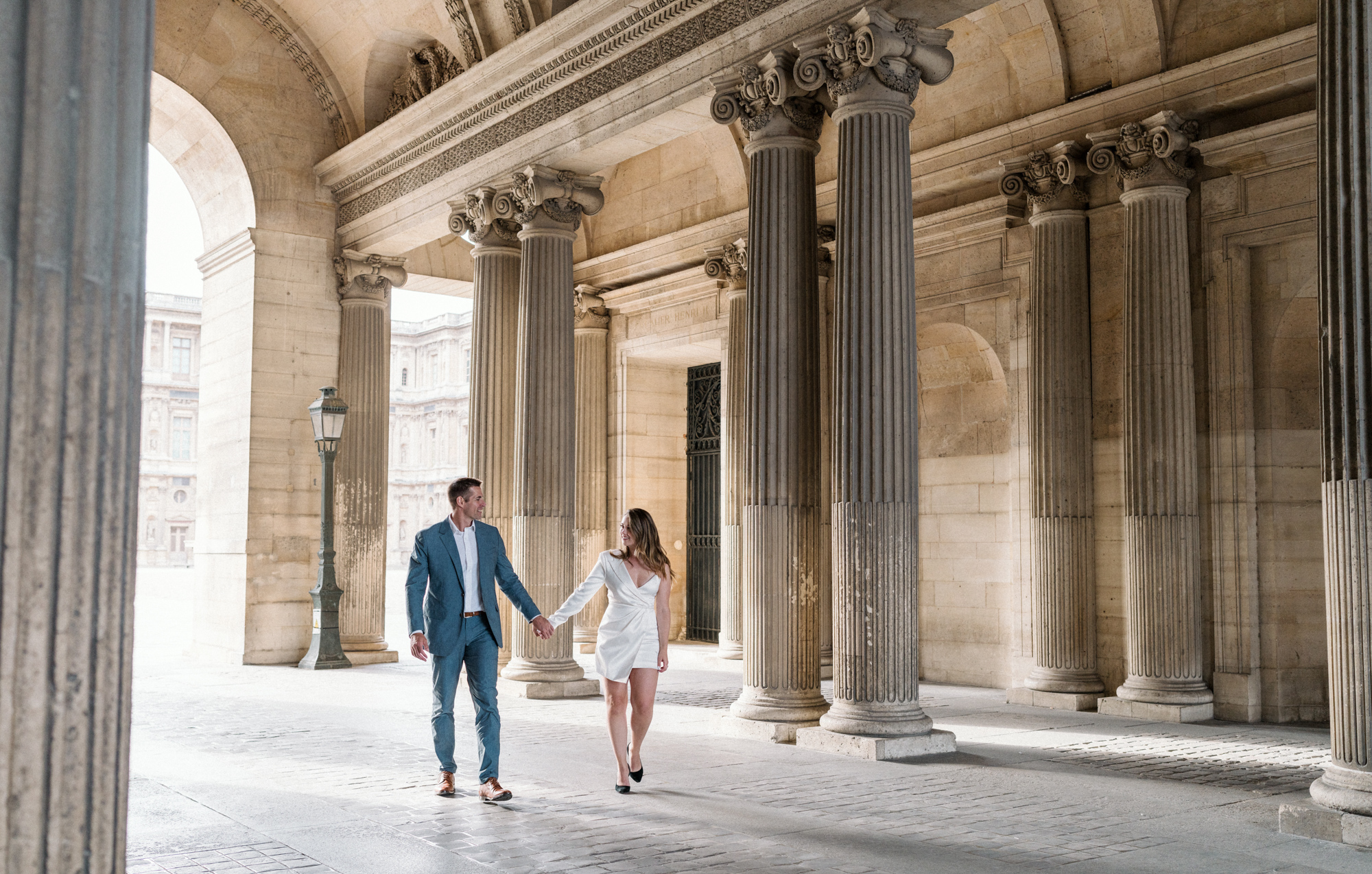 engaged couple walk hand in hand smiling at louvre in paris