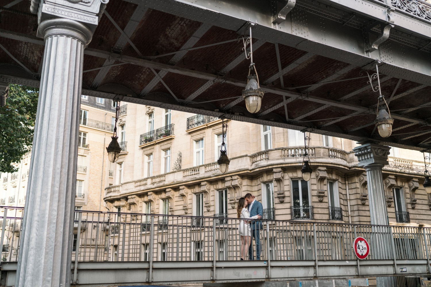 engaged couple embrace on bridge with view of paris architecture