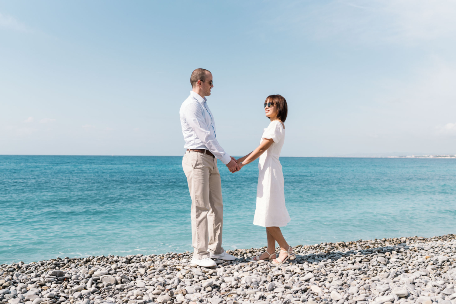 man and woman wearing sunglasses pose on stone beach with blue water in background