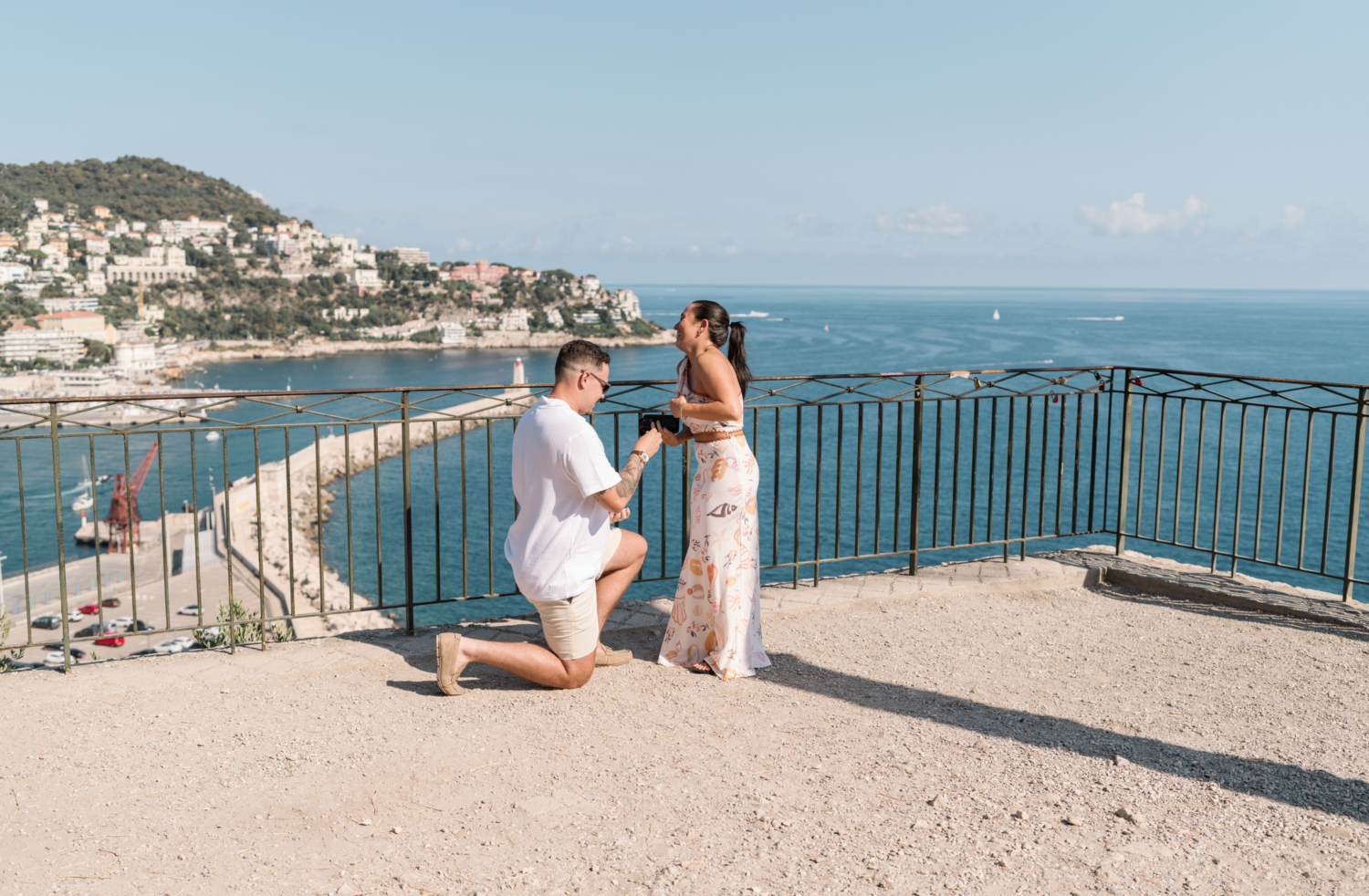 woman laughs with surprise as man proposes to her with view of sea in nice france