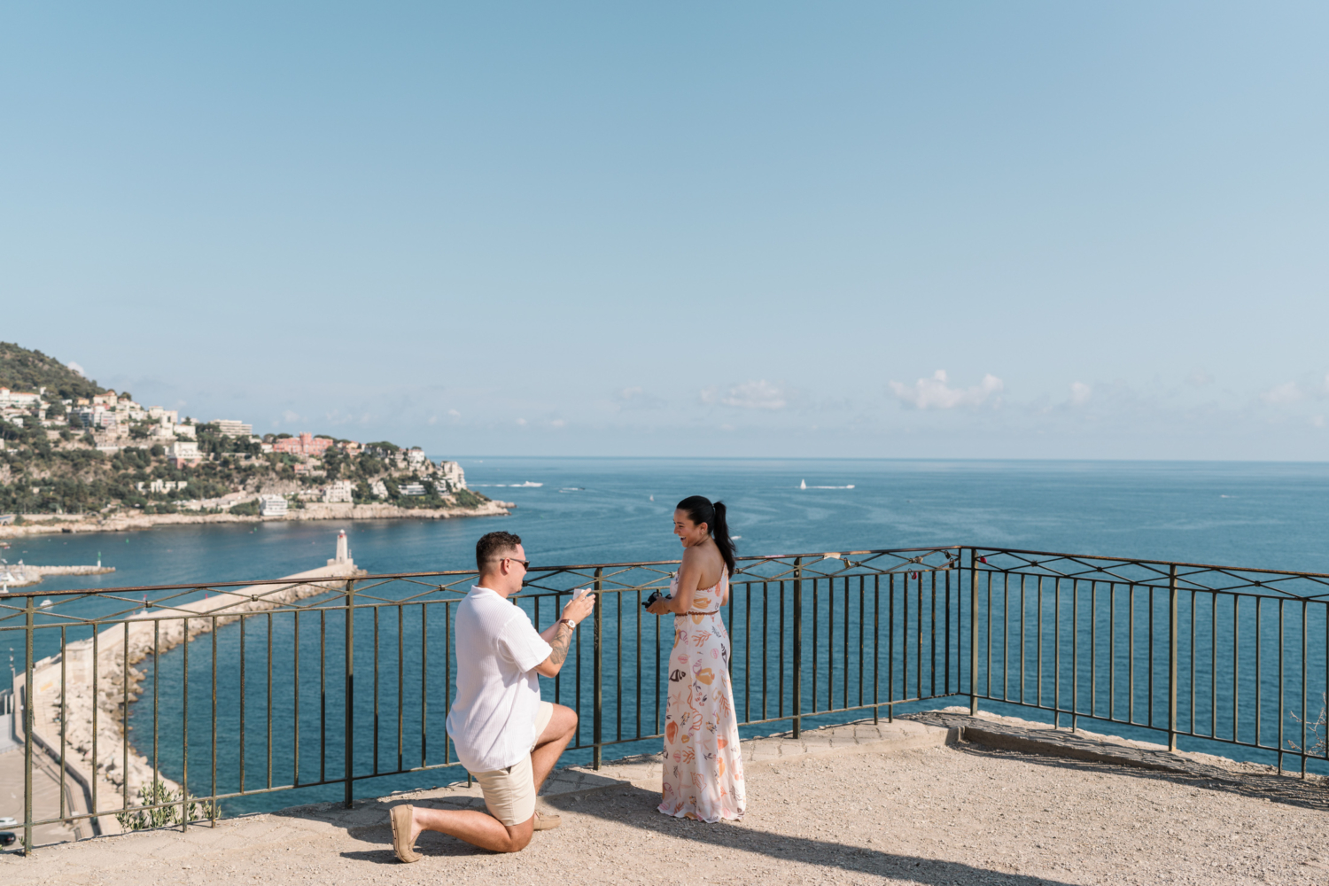 woman laughs as man gets on one knee to propose with view of sea in nice france