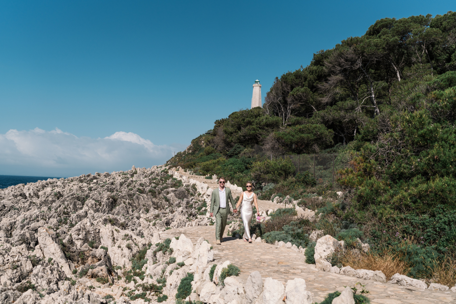 bride and groom walk down path with lighthouse behind in saint jean cap ferrat