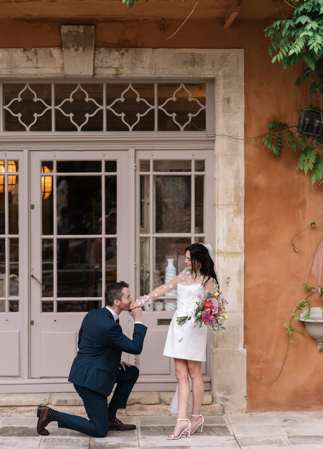 groom kisses hand of bride next to colorful building in gordes france