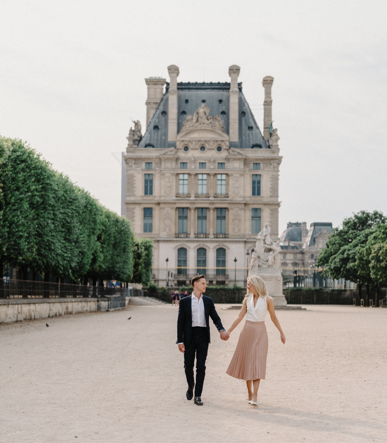engaged couple walk hand in hand with view of louvre in paris