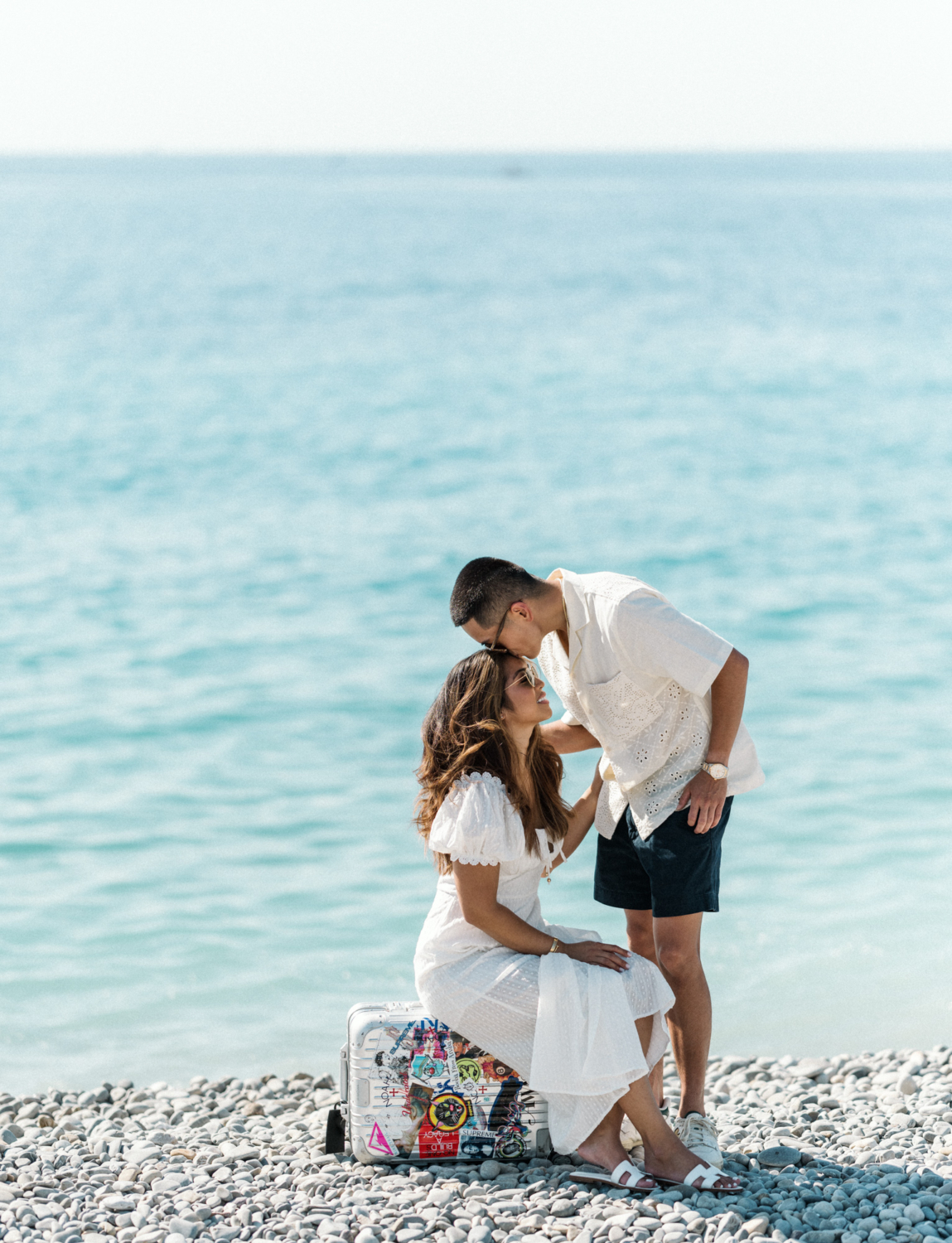 woman sits on suitcase while man kisses her head on beach in nice france