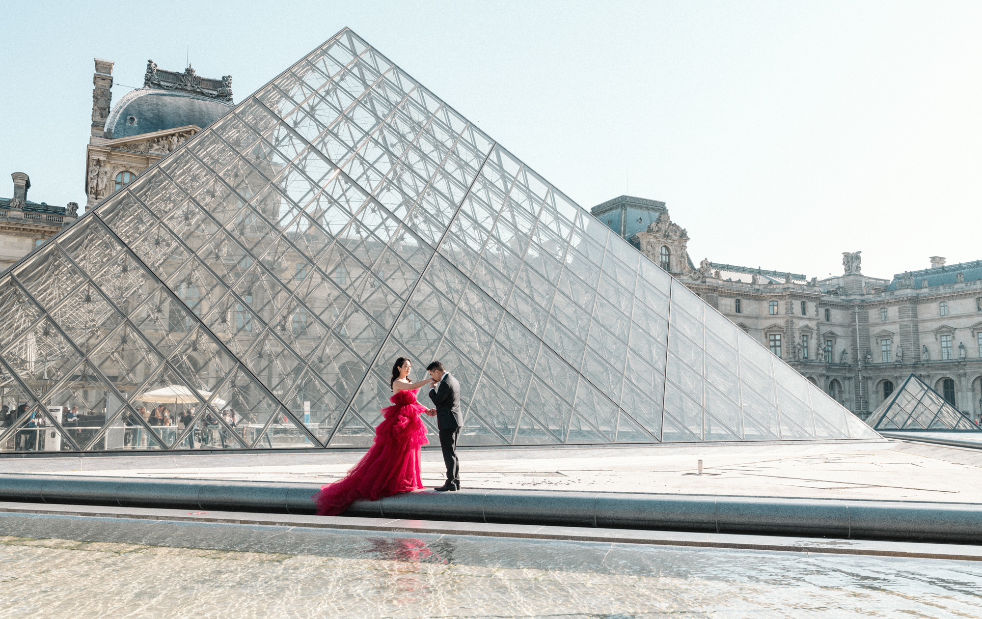 asian man kisses hand of asian woman in front of glass pyramid at louvre in paris