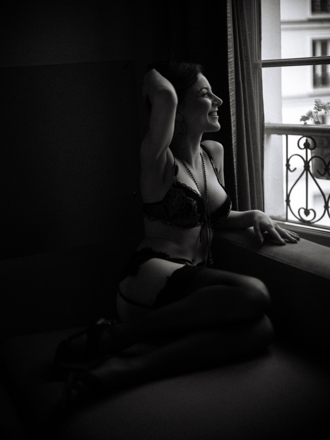 woman in lingerie smiles and looks out window during boudoir photoshoot in paris