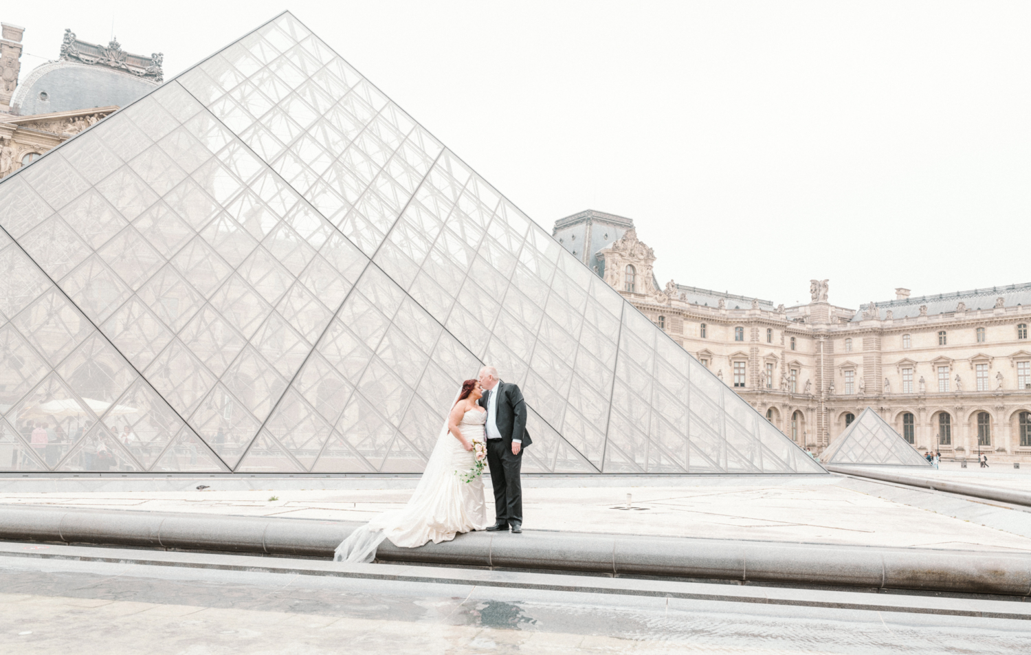 groom kisses bride's forehead at glass pyramid in paris