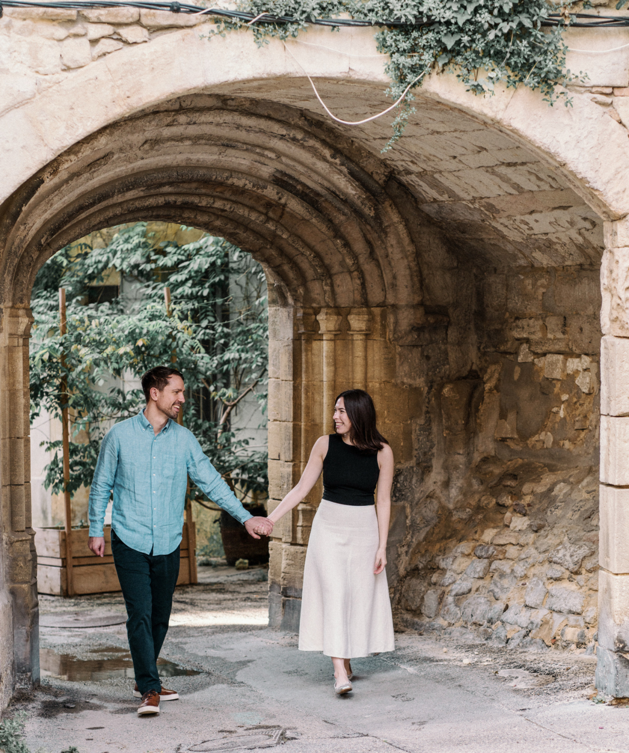 smiling engaged couple walk hand in hand through archway in arles