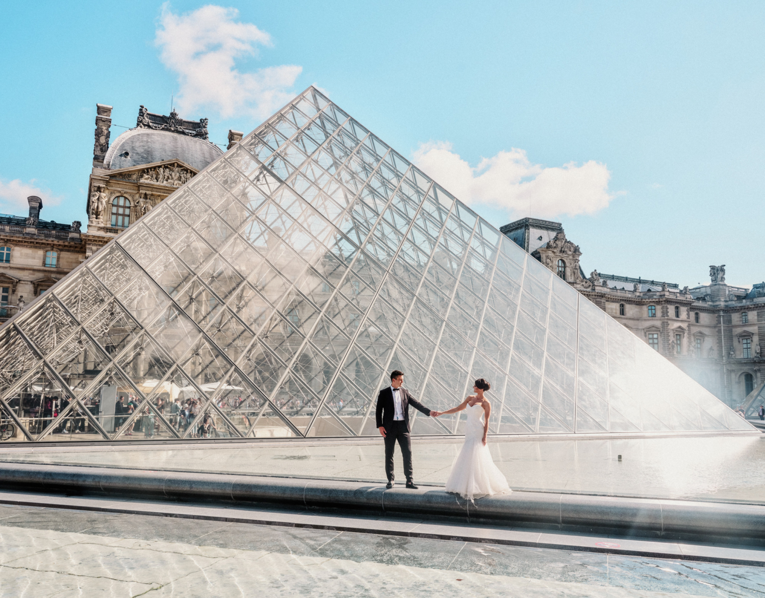 bride and groom pose in front of glass pyramid at louvre in paris