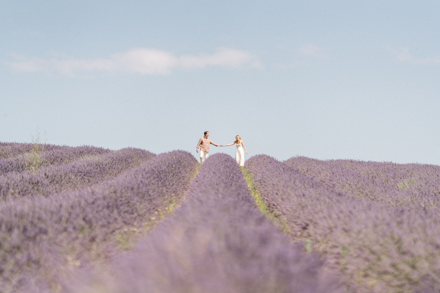 man and woman hold hands in lavender field with dreamy blue sky