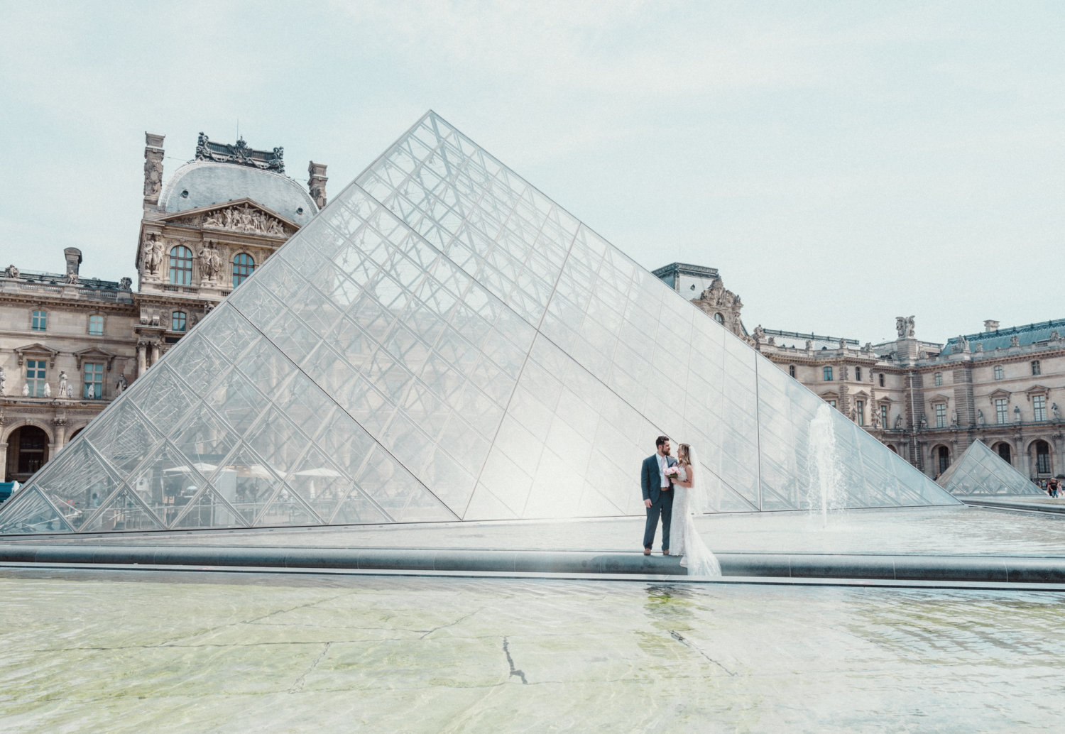 bride and groom pose in front of glass pyramid at louvre in paris