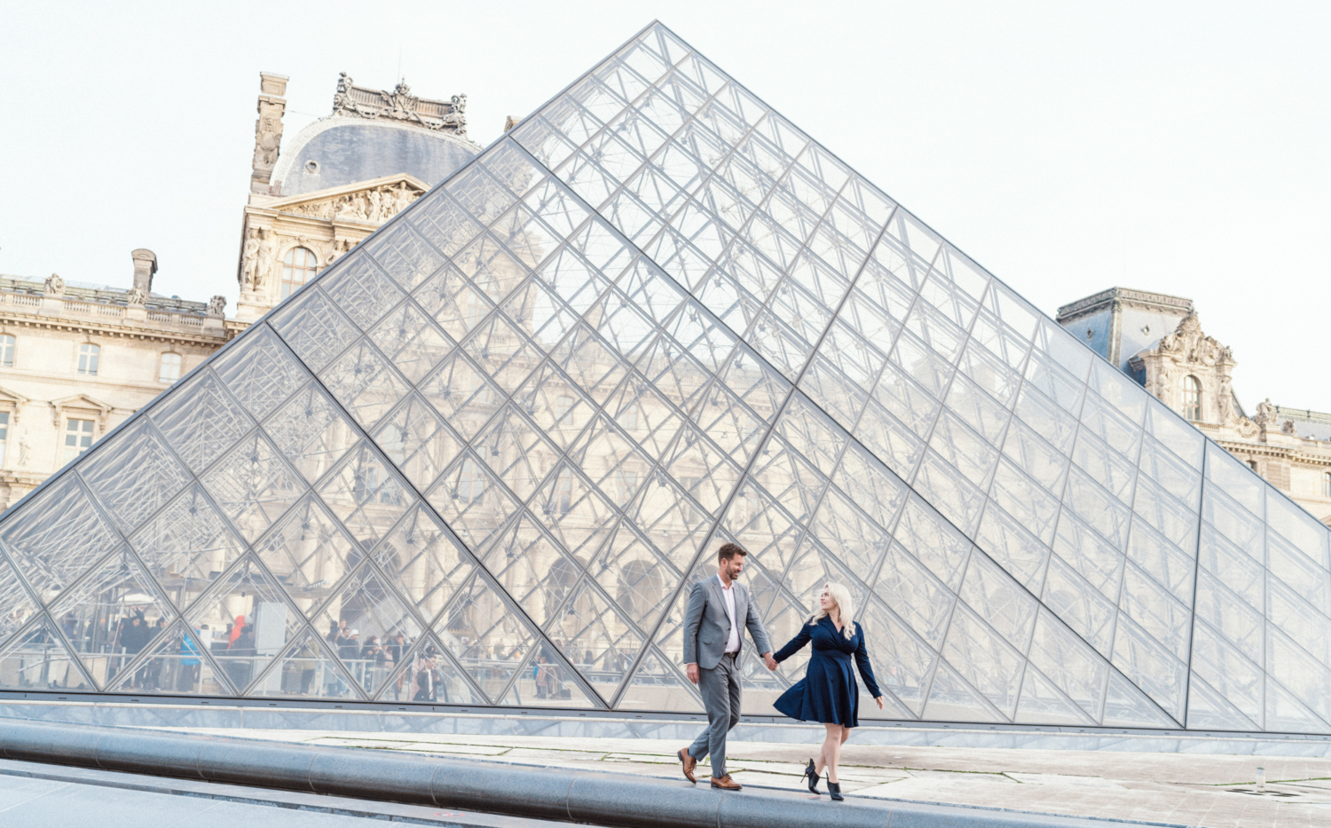 couple walk in front of glass pyramid at louvre in paris