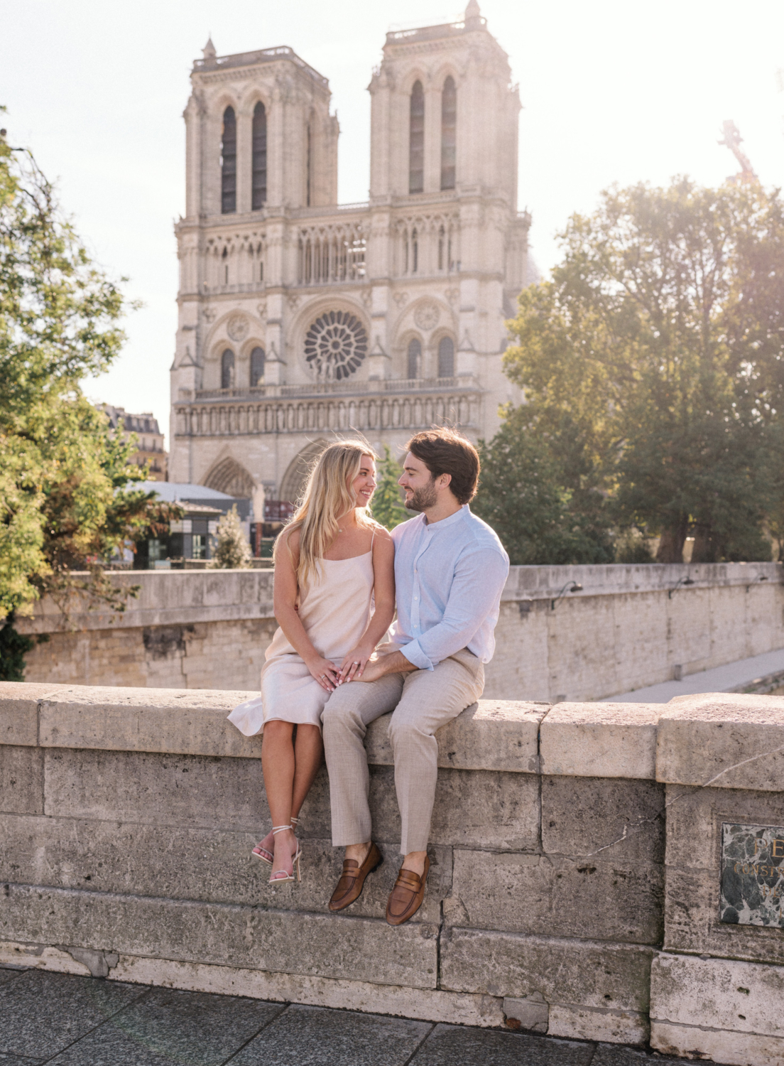 engaged young couple sit on wall with view of notre dame cathedral in paris