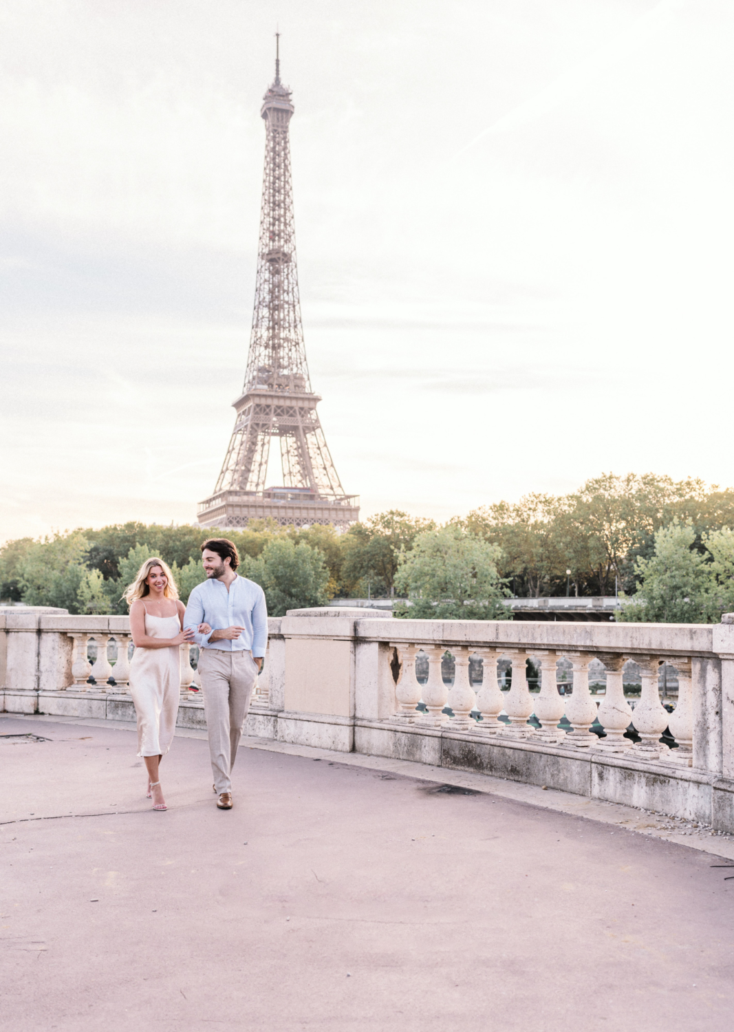 engaged young couple laugh while walking arm in arm with view of eiffel tower