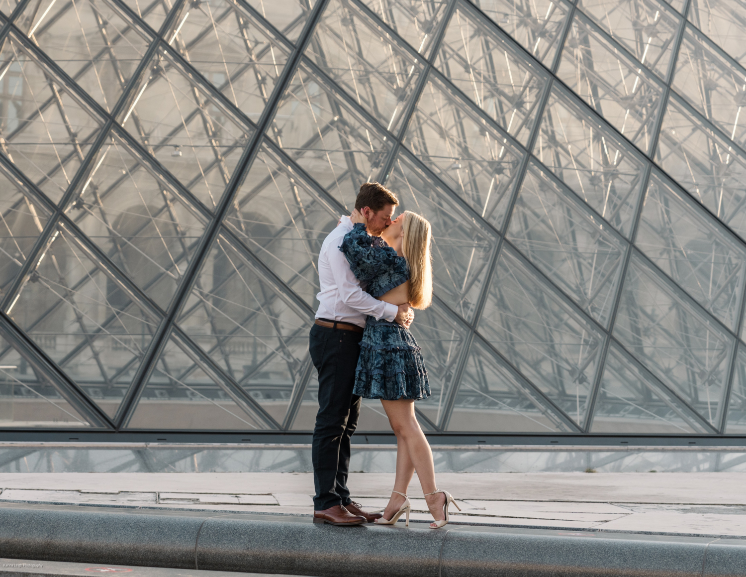 young newly engaged couple kiss at the glass pyramid at the louvre museum in paris