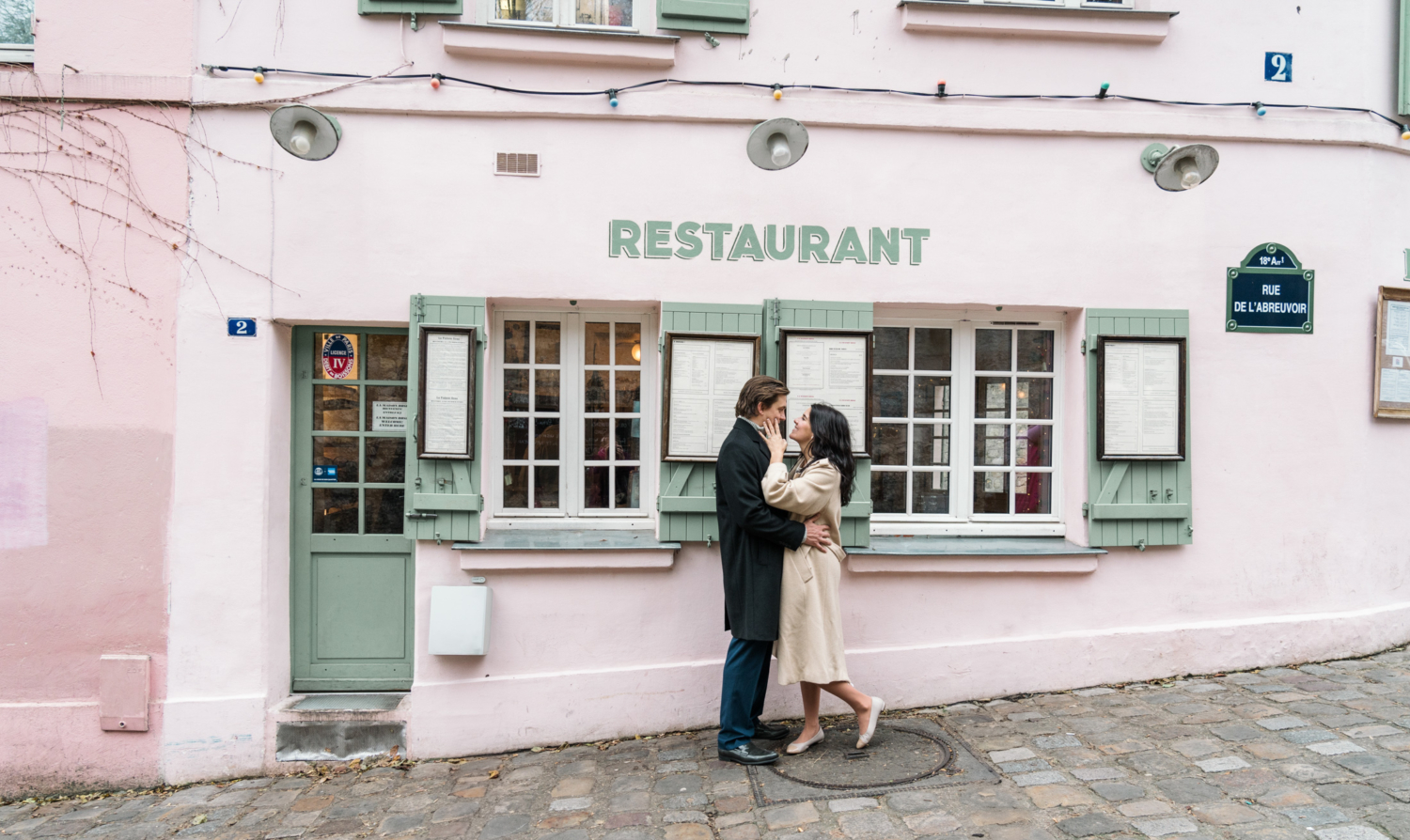 newly engaged couple embrace in front of charming pink restaurant in montmartre during Engagement Photos In Paris