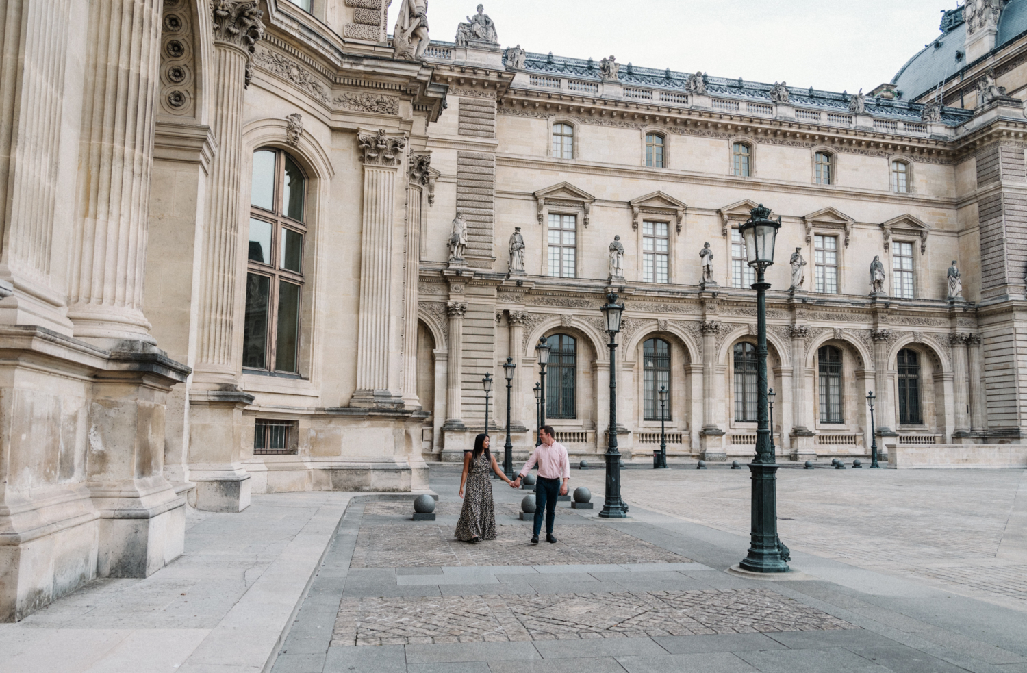 newly engaged couple hold hands as they walk through the louvre courtyard in paris