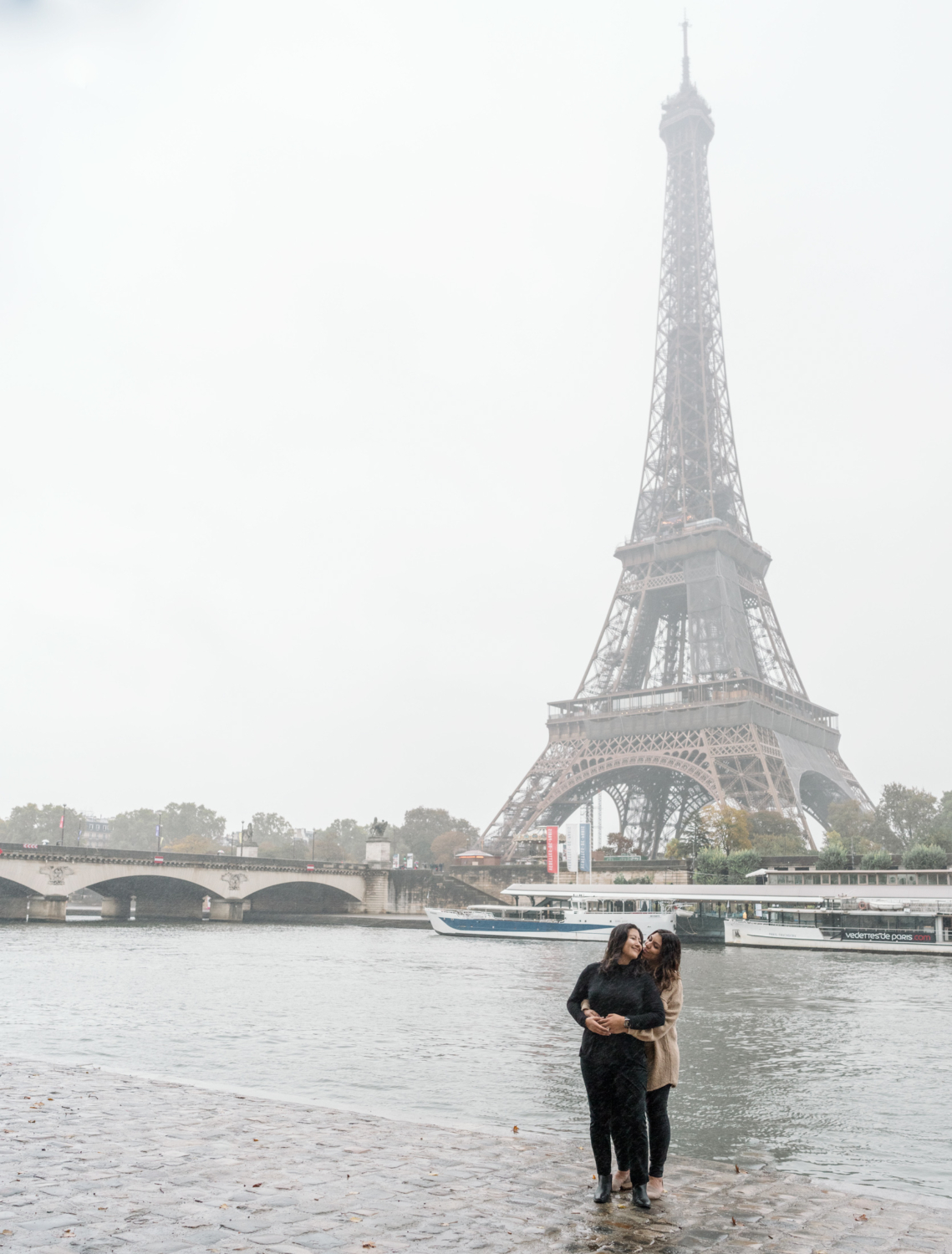 two women embrace and kiss with view of the eiffel tower in background in paris