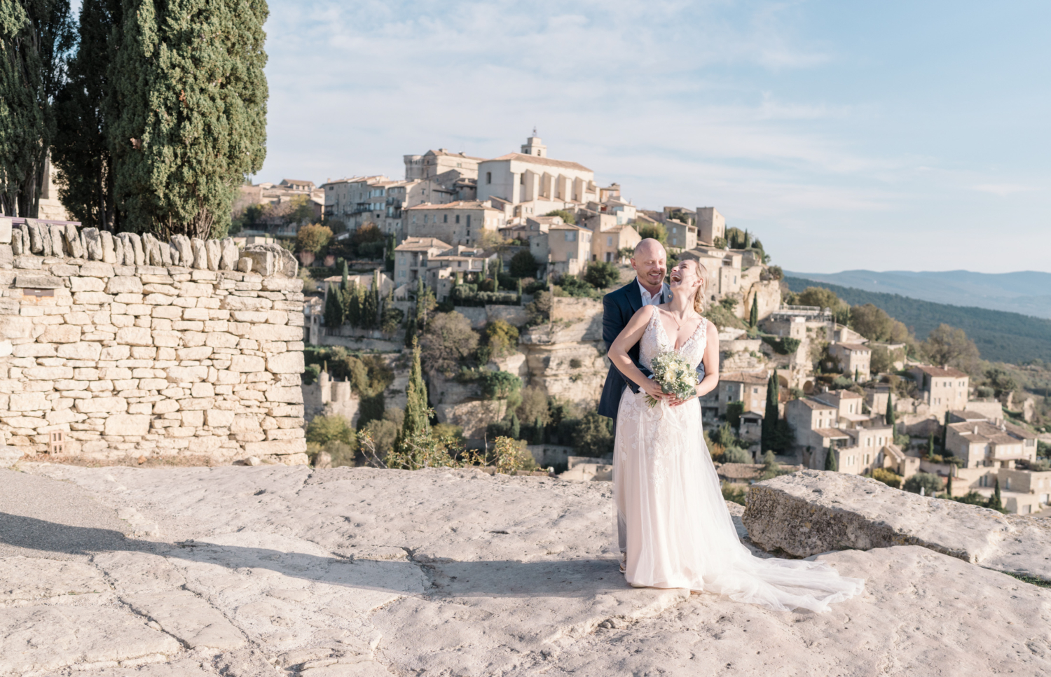 bride and groom laugh with village of gordes france provence in background