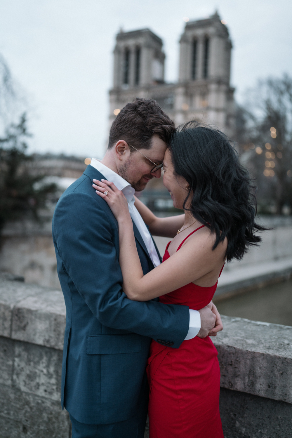 newly engaged couple smile and embrace with view of notre dame cathedral in paris