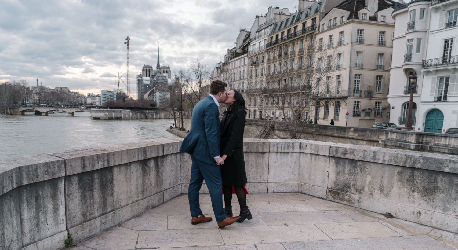 newly engaged couple kiss after their surprise proposal with view of notre dame in paris