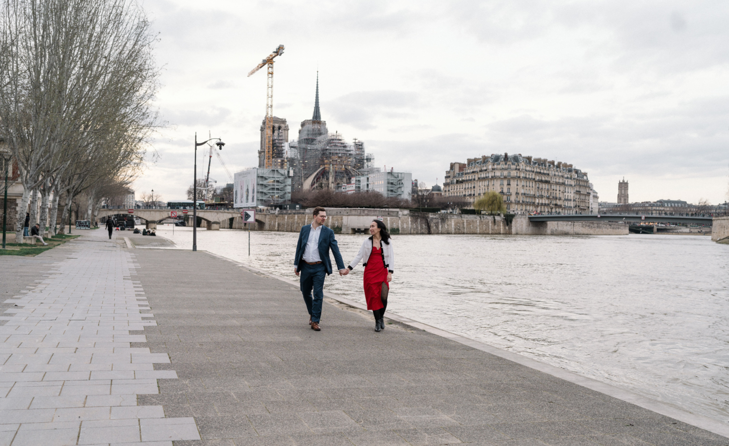 newly engaged couple walk hand in hand in paris with view of notre dame cathedral