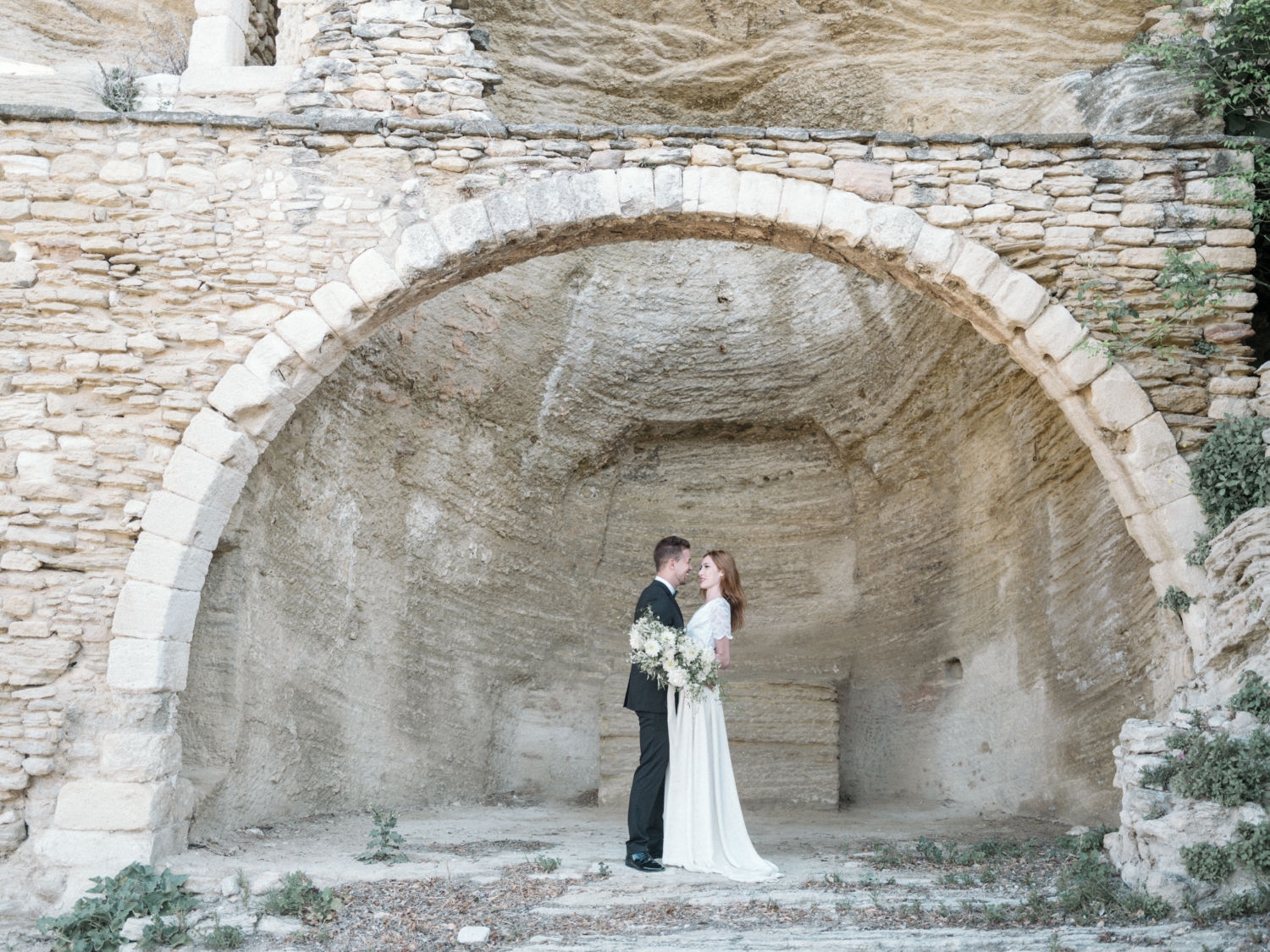 bride and groom pose under stone arch in gordes france during their La Bastide de Gordes wedding