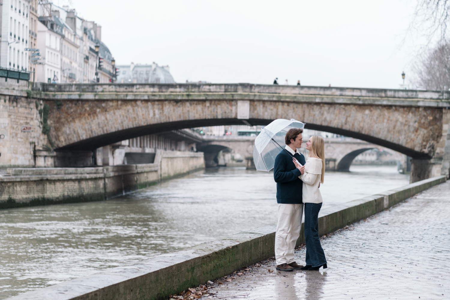 newly engaged couple embrace in the rain along the river in paris