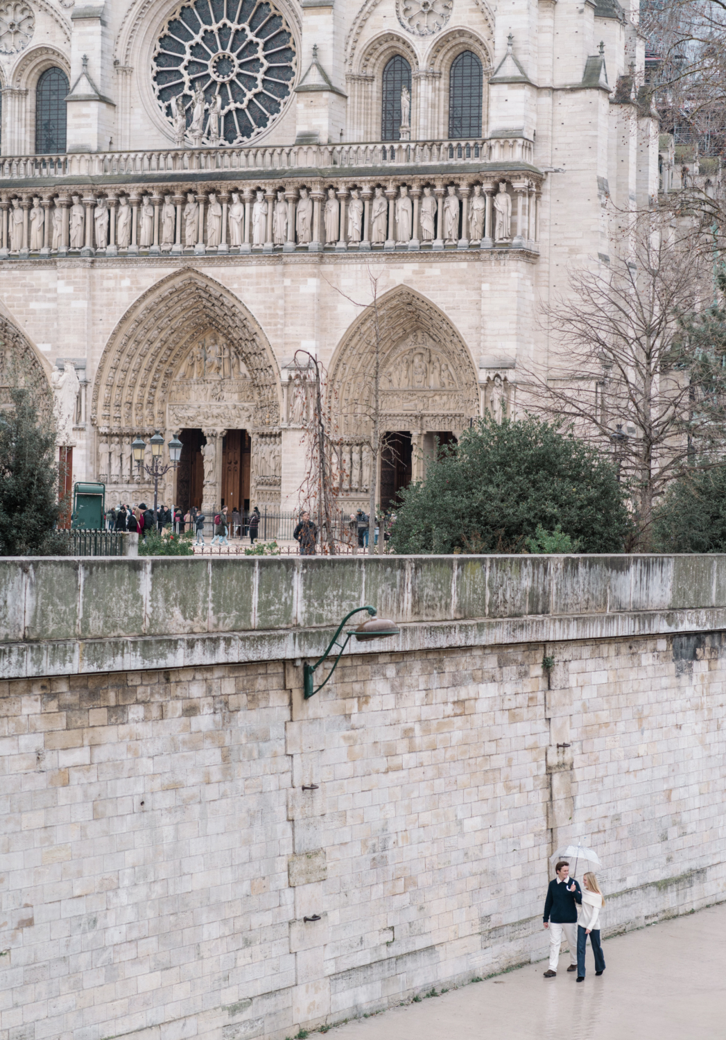 newly engaged couple walk along the river in paris with notre dame cathedral in the background