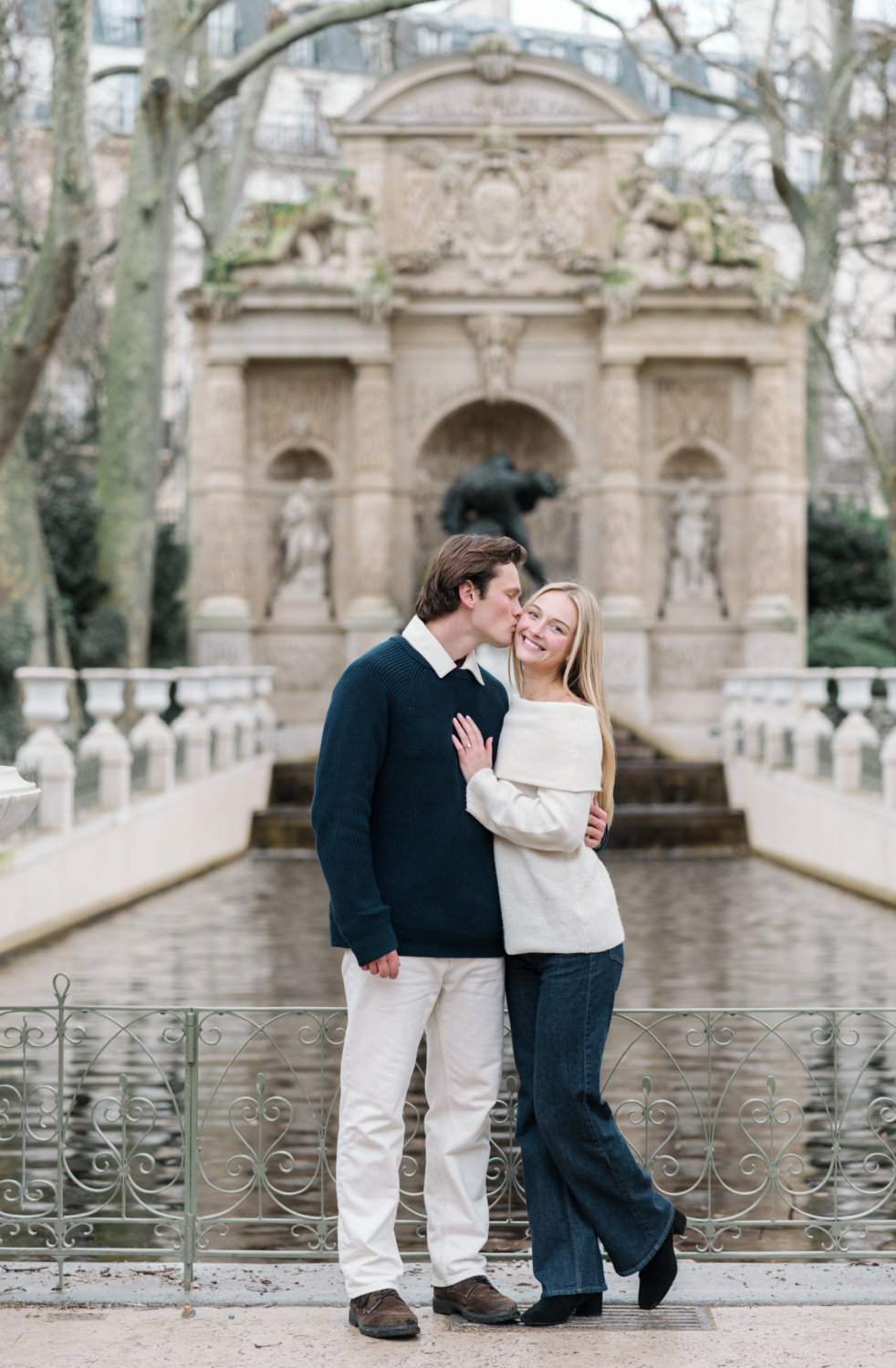 man kisses woman on cheek in front of fountain in luxembourg gardens in paris