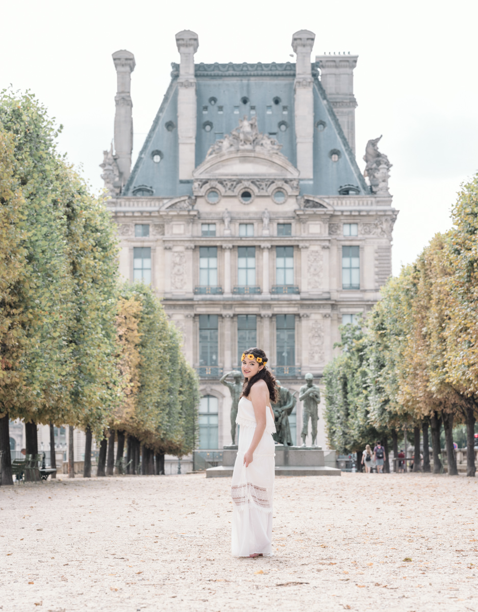 young woman in white dress wearing a flower crown smiles in the tuileries gardens in paris