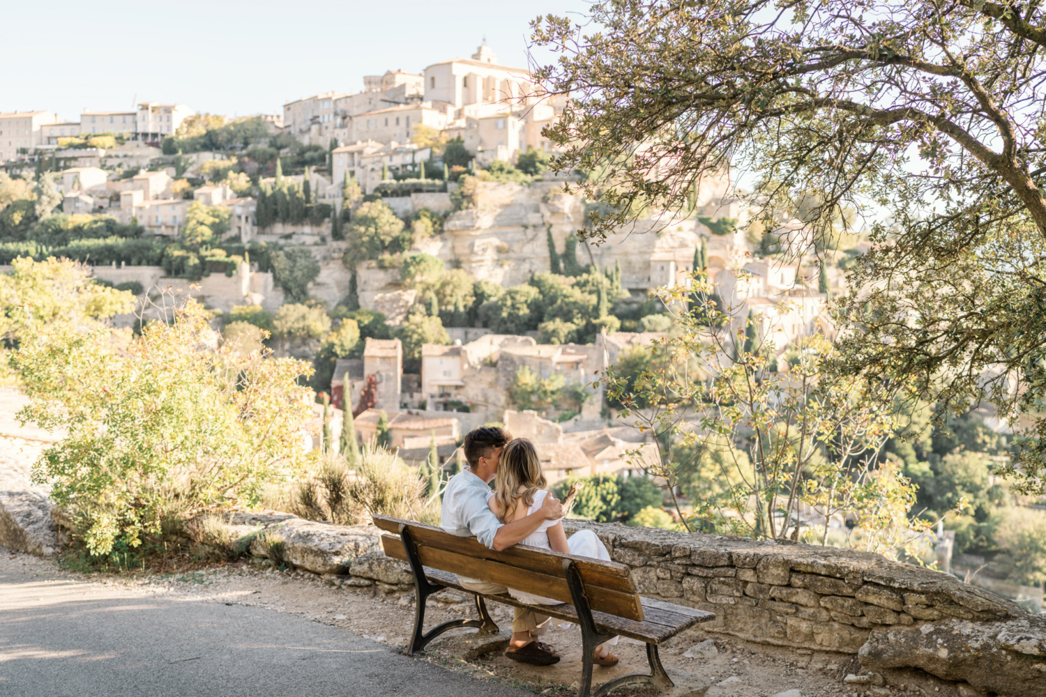 engaged couple sit on a bench overlooking gordes, provence france